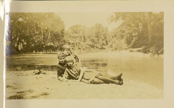 A faded sepia-toned photograph of four White women, all wearing headwraps, posing on the ground next to a river.