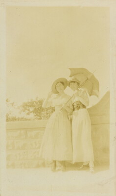 A faded sepia-toned photograph of two White women in sunhats and long skirts, one holding an open parasol, standing next to a stone wall.