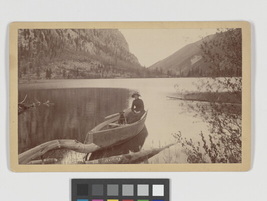 A sepia-toned photograph of a woman and a dog in a canoe on a still lake in the mountains.