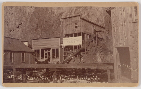 A black-and-white photograph of wood buildings built into the side of a steep cliff.