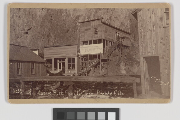 A black-and-white photograph of wood buildings built into the side of a steep cliff.