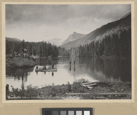 A black-and-white photograph of people canoeing and fishing on a lake surrounded by trees and mountains.