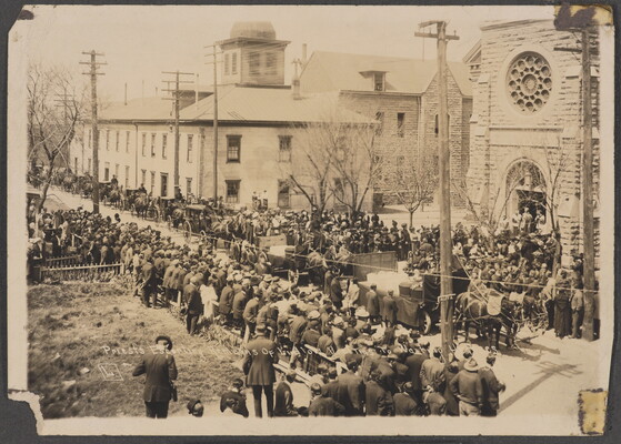 A black-and-white photograph of crowds lining a street outside a of church watching as coffins are loaded onto horse-drawn carriages.