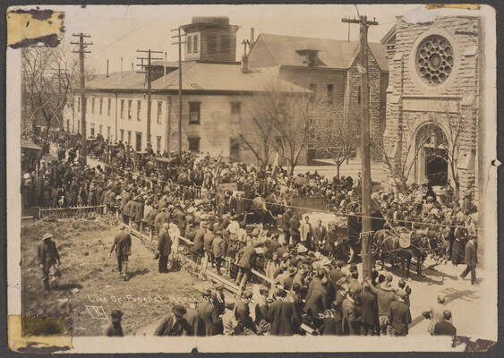 A sepia-toned photograph of crowds lining a street outside a of church watching as coffins are loaded onto horse-drawn carriages.