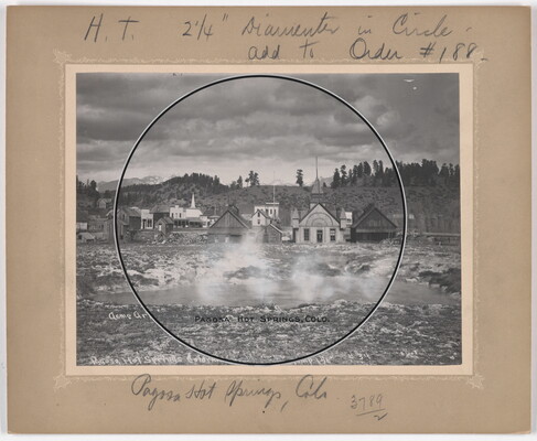 A black-and-white photograph of a hot spring with steam rising off the water and houses and hills in the background.