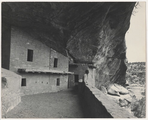 A black-and-white photograph of a man standing in a stone cliff dwelling overlooking the valley beyond.