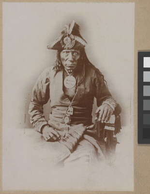 A black-and-white portrait photograph of a seated Indigenous man seated wearing a colonial-style jacket and a tricorn hat.