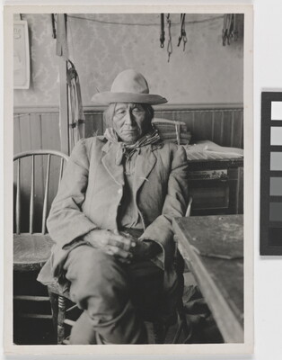 A black-and-white photograph of an Indigenous man in a rumpled suit seated next to a table with his hands in his lap.