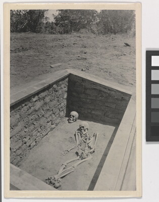 A black-and-white photograph of an archaeological grave site with a full human skeleton laying beside a skull.