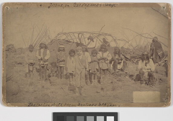 A slightly faded sepia-toned photograph of a group of Indigenous adults and children and one White child standing in front of a camp.