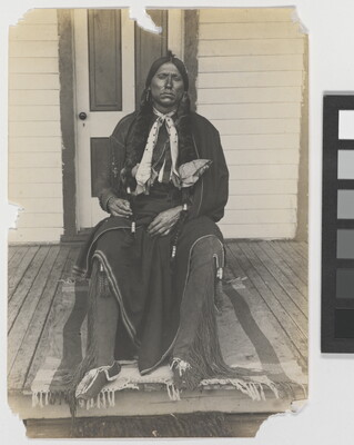 A black-and-white photograph of an Indigenous man with long braids seated on a wooden porch.