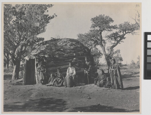 A black-and-white photograph of a group of Indigenous adults and children in front of a round, wood dwelling.