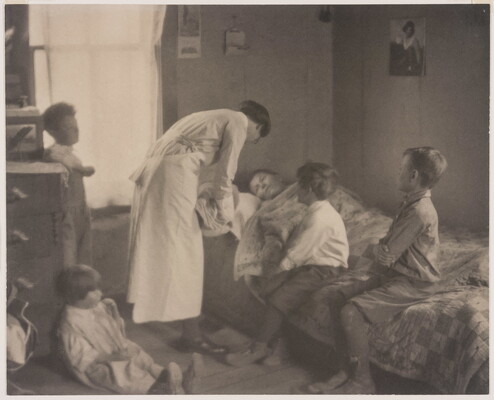 A black-and-white photograph of a nurse bending over a person in a bed, two children sitting on the bed, and two children behind the nurse.