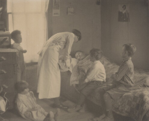 A black-and-white photograph of a nurse bending over a person in a bed, two children sitting on the bed, and two children behind the nurse.