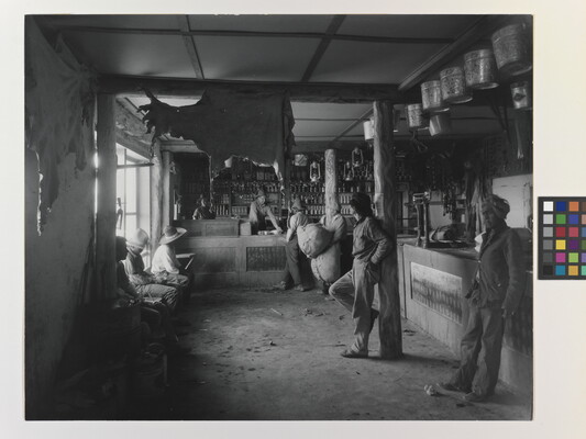 A black-and-white photograph of Indigenous people in a shop with a variety of products such as animal hides, tin pails, and lanterns hanging from the ceiling.