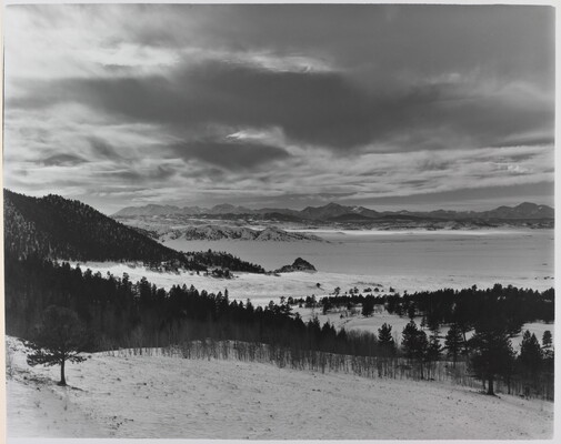 A black-and-white photograph of a snowy mountain plain with tree-covered hills and a cloudy, but illuminated, sky.