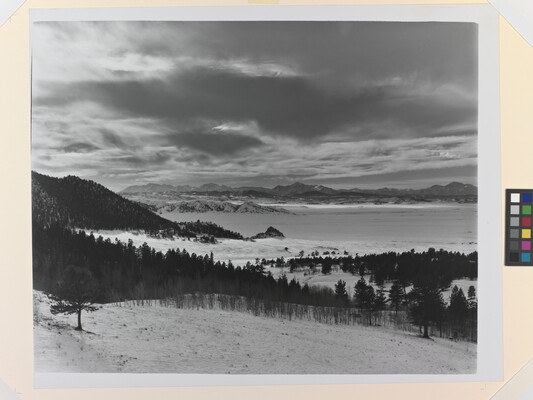 A black-and-white photograph of a snowy mountain plain with tree-covered hills and a cloudy, but illuminated, sky.