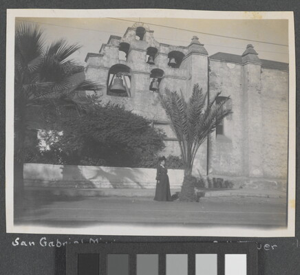 A black-and-white photograph of a woman standing next to a building with several large bells hanging near the top.