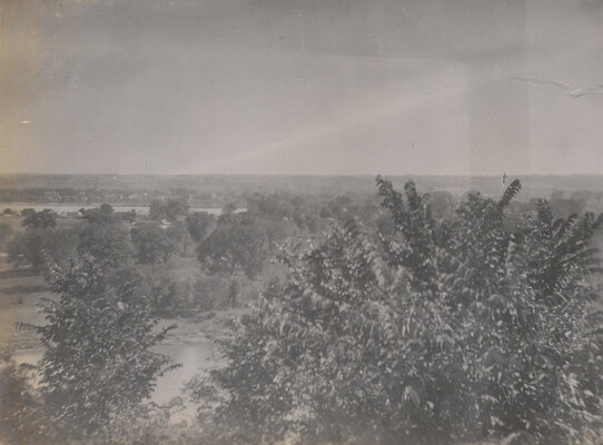 A black-and-white photograph taken from a higher elevation of a forested area and a river in the distance.