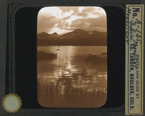 A black-and-white image of light reflected on a body of water under a cloudy sky with mountains in the distance.