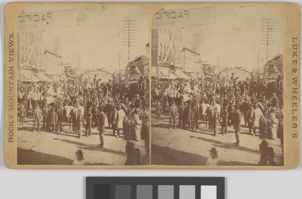 A sepia-toned stereograph of a large gathering of people in a street lined with buildings.