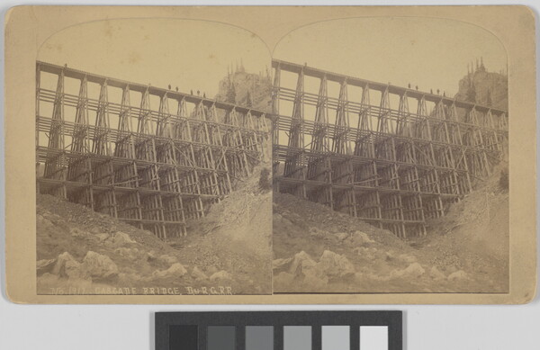 A sepia-toned stereograph of a high trestle bridge over a mountainous ravine viewed from the bottom.