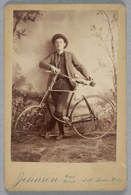 A black-and-white studio photograph of a young White man wearing a hat and riding clothes posing with a bicycle in front of him.