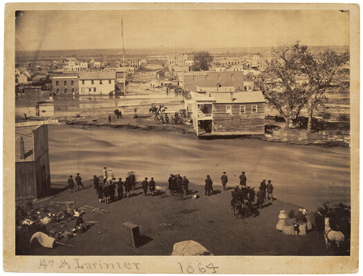 A sepia-toned photograph of damaged buildings and streets surrounded by mud and water as people look at the devastation.