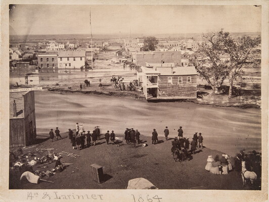 A sepia-toned photograph of damaged buildings and streets surrounded by mud and water as people look at the devastation.