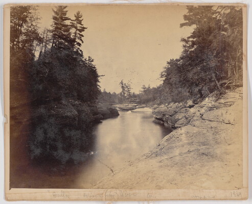 A sepia-toned photograph of a river with rock formations on one side and tall trees on the other.