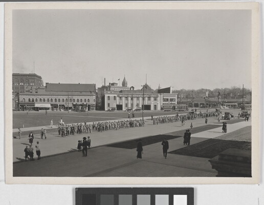 A black-and-white photograph of a parade coming through a wide street as scattered spectators watch.