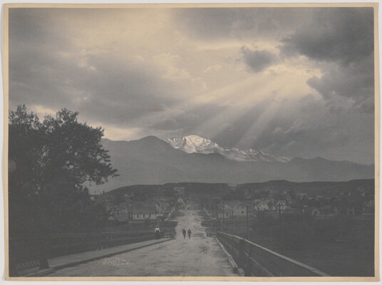 A black-and-white photograph of two people walking down a road toward a town; snow-capped mountain and sun rays in the distance.