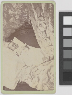A black-and-white photograph of a White baby wearing a white gown and positioned on a pillow framed by a floral curtain.