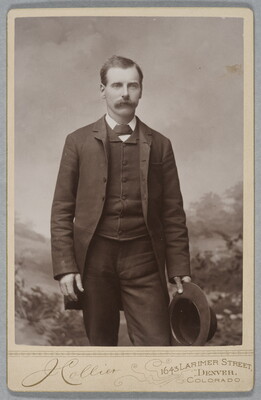 A black-and-white studio photograph of a young White man with a mustache wearing a three piece suit, holding a hat in one hand.