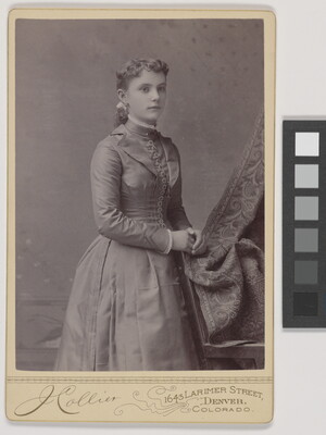 A black-and-white studio photograph of a young White woman in a fitted, long-sleeved dress, hands resting on the back of a chair.