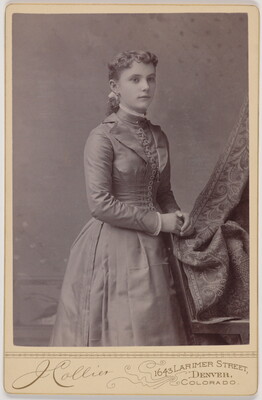 A black-and-white studio photograph of a young White woman in a fitted, long-sleeved dress, hands resting on the back of a chair.