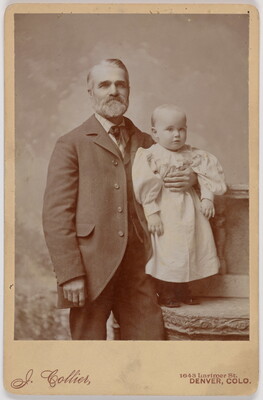 A black-and-white studio photograph of a White man with graying hair and beard wearing a suit with his arm around the torso of a child who is standing on a chair and wearing a long-sleeved dress.