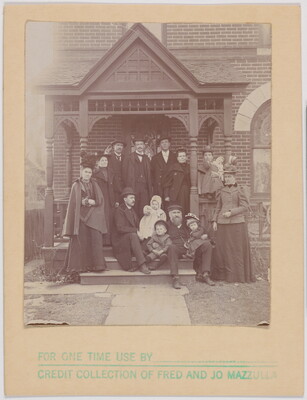 A black-and-white photograph of a well-dressed White family posing on the front steps of a brick Victorian home.