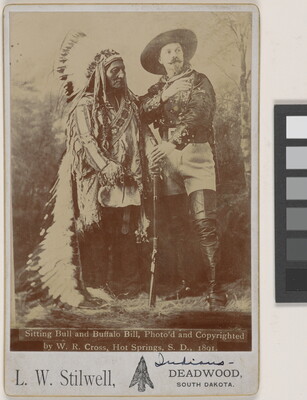 A sepia-toned portrait photograph of an Indigenous man wearing a feathered headdress and traditional clothing next to a White man in a large hat and tall leather boots holding a rifle in front of them.
