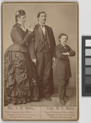A sepia-toned portrait photography of a very tall, White woman in an elaborately draped dress next to a very tall, White man, and a White man of average height, both in suits.