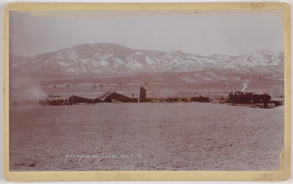 A black-and-white photograph of mangled train cars and an engine on a plain with snow-covered mountains in the distance.