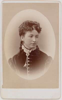A black-and-white oval-shaped portrait photograph of a White woman, hair pulled up with ringlets that lay on her shoulders, wearing a dress with a high ruffled neck.