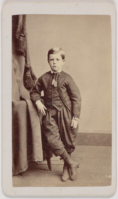 A black-and-white portrait photograph of a White boy wearing a formal jacket and vest over knickerbocker pants, leaning on a table, one leg crossed over the other.