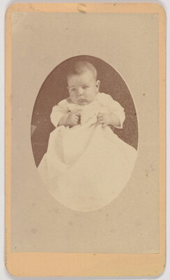 A black-and-white oval-shaped portrait photograph of a White infant in a white gown.