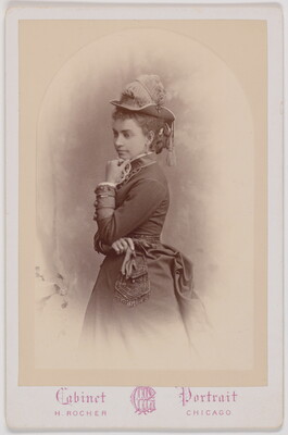 A black-and-white portrait photograph of a young White woman, standing in profile, wearing a hat decorated with feathers and tassels, holding a beaded purse in one hand, the other hand at her chin.