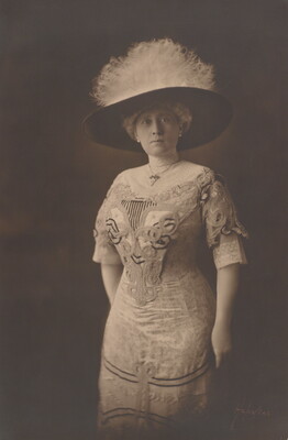 A black-and-white photograph of a well-dressed White woman in a large feathered hat and ornate beaded dress.