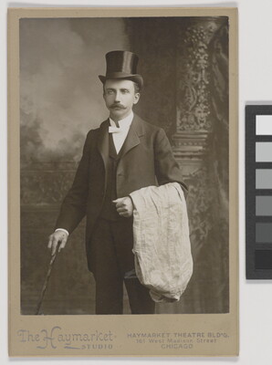 A black-and-white studio photograph of a White man with a mustache, wearing a top hat and three-piece suit, holding a cane in one hand, white cloth draped over the other arm.