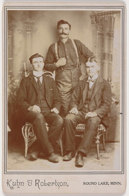A black-and-white studio photograph of three White men: one standing wearing pants with suspenders; two seated, both wearing three-piece suits.
