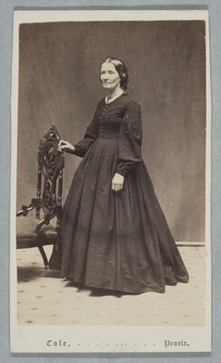 A black-and-white studio photograph of a middle-aged White woman wearing a dark long-sleeved dress with a full skirt resting one hand on the back of a chair.