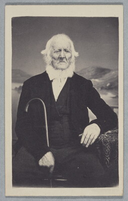 A black-and-white studio photograph of a seated elderly White man with white hair and beard, wearing a black suit, holding a cane in one hand.